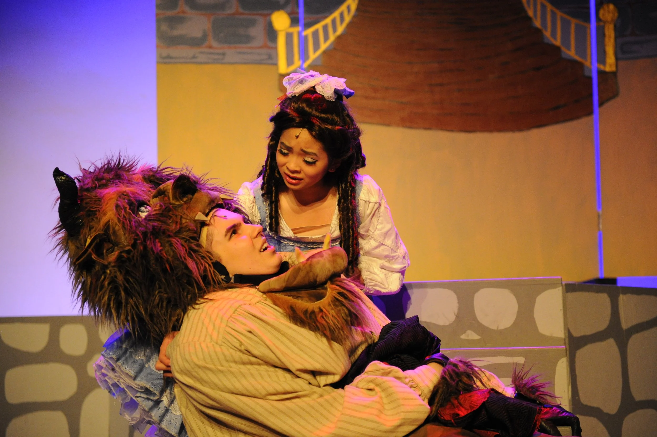 Two actors on stage, one wearing a lion costume and lying down, the other dressed as a girl with braids and a bow, crouching beside the lion and looking at it, with a painted backdrop of a brick wall and a staircase.