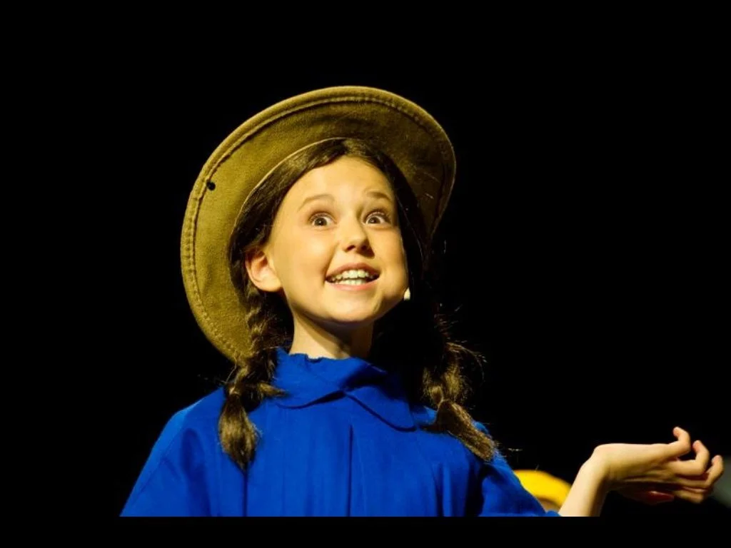 A young girl dressed as Dorothy from The Wizard of Oz, wearing a blue dress and a straw hat, smiling and gesturing with her hand against a black background.