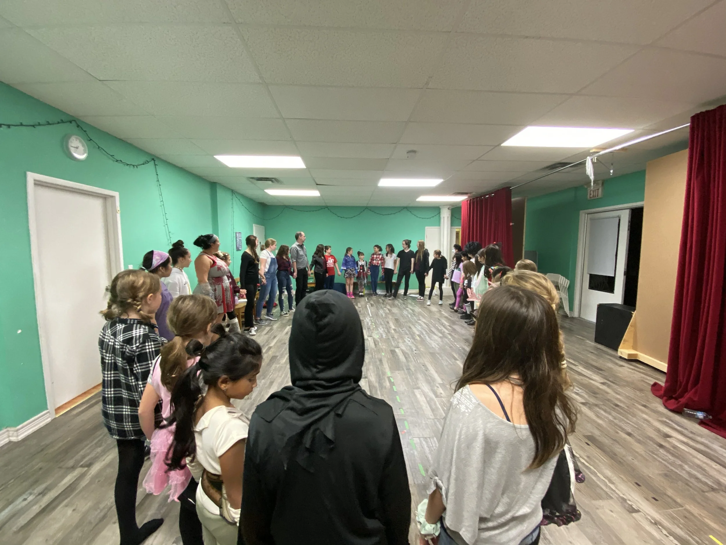A group of children and adults standing in a circle in a room with teal walls, red curtains, and wood flooring, possibly participating in a dance or game.