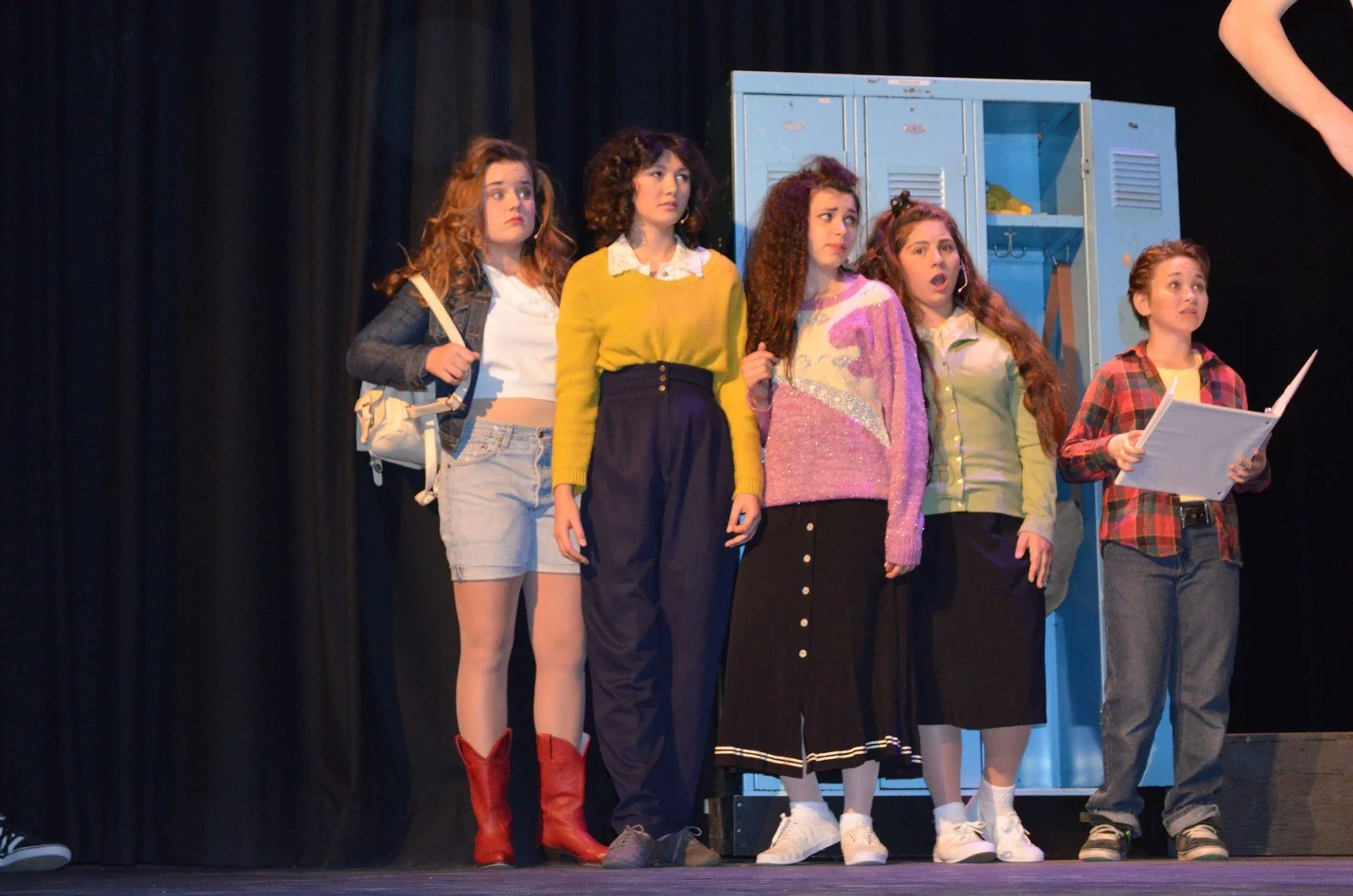 Five young girls standing on a stage, possibly in a school play, with a blue locker in the background. One girl on the far right is holding a script or booklet.