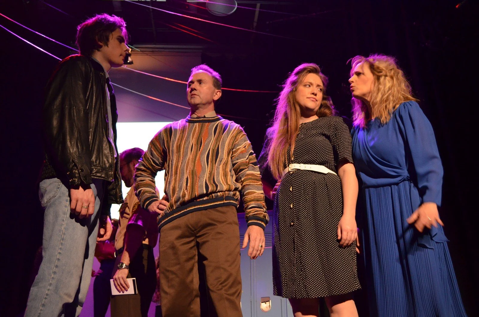 Four people on stage engaged in conversation. The person on the left is a young man with dark hair wearing a leather jacket and jeans. The second person is an older man with gray hair in a colorful striped sweater. The third person is a young woman w