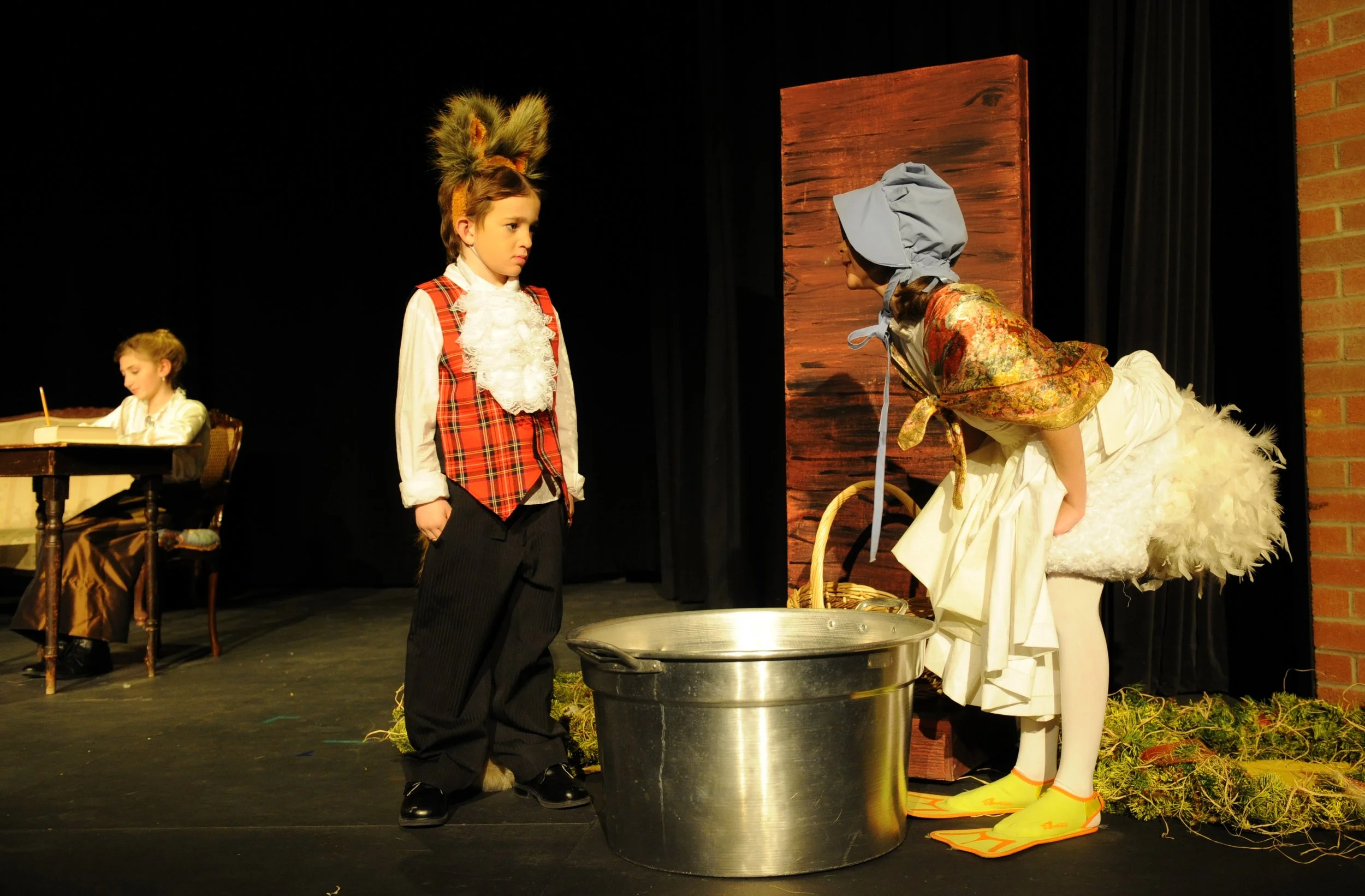 Two children performing in a play. One girl wearing a maid costume and a bonnet, and a boy dressed as a character with a wig and fancy vest. In the background, a girl seated at a table with a book, and a woman dressed as a goose with a headscarf, apr