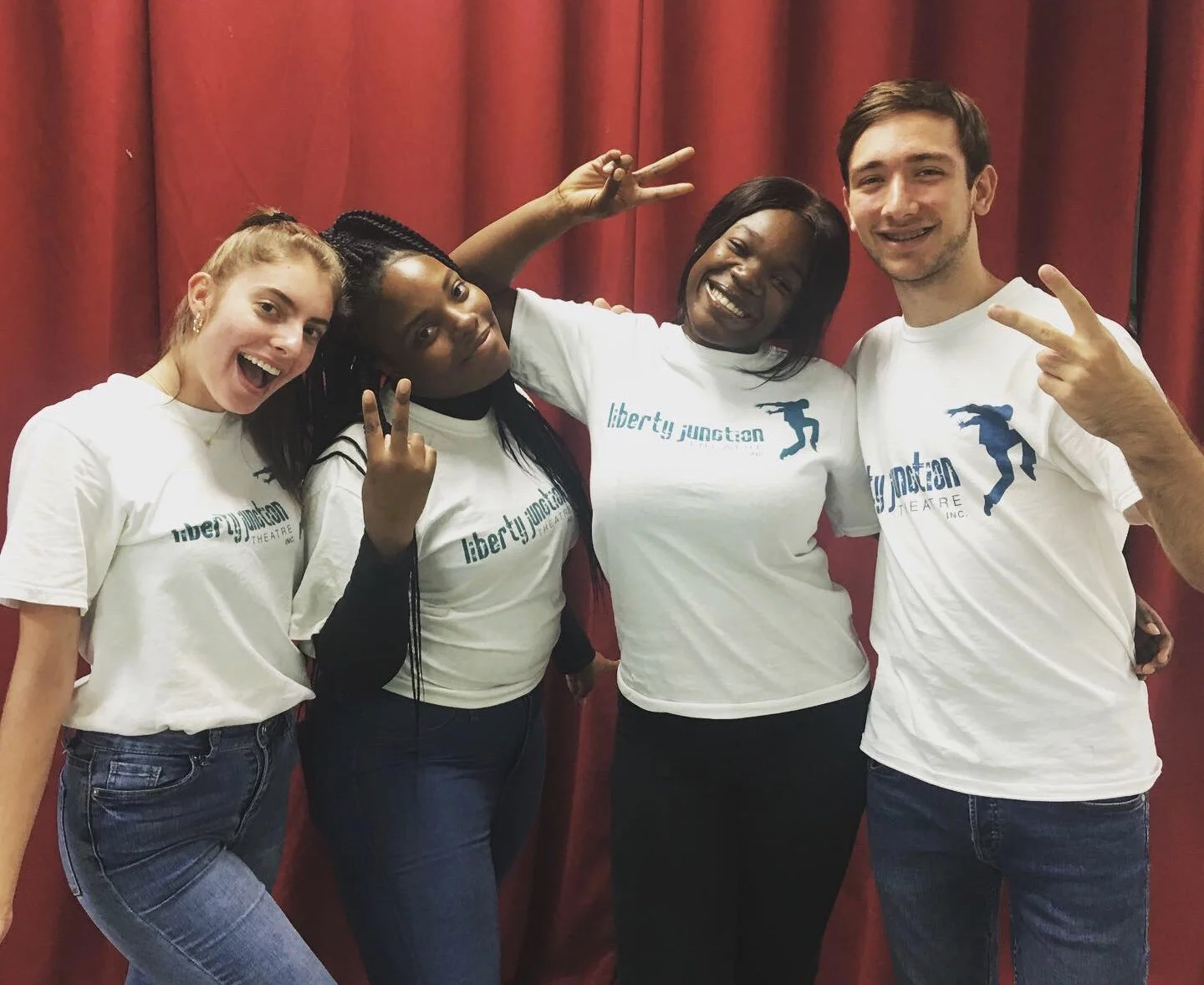 Group of four diverse young adults wearing white t-shirts with 'liberty junction' logo, smiling and posing with peace signs in front of a red curtain.