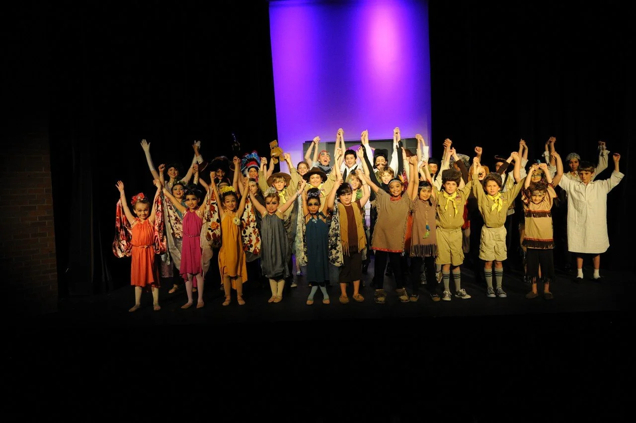 Children on stage dressed in costumes, raising their arms in a group celebration after a performance.