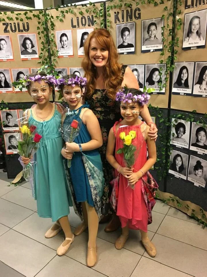 Three young girls dressed in colorful ballet costumes with flower crowns and holding flowers, standing beside a smiling woman with long red hair in front of a backdrop featuring black and white photos of children and the words "Zulu Lion King".