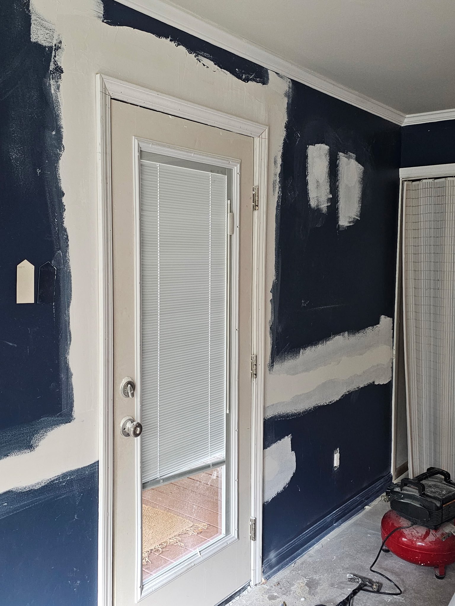 Interior wall near a door with black paint in progress, partially painted over white wall. A window with blinds on the door and a screen door to the outside. Tools and workshop items on the floor including a red shop vacuum.