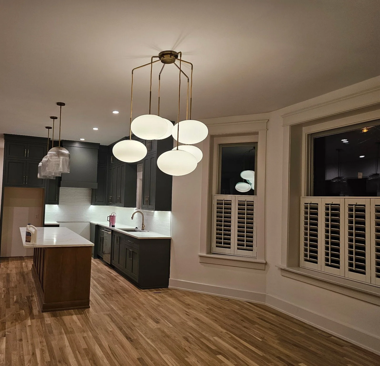 Modern kitchen with dark gray cabinets, white walls, wooden flooring, and stylish lighting fixtures hanging from the ceiling.