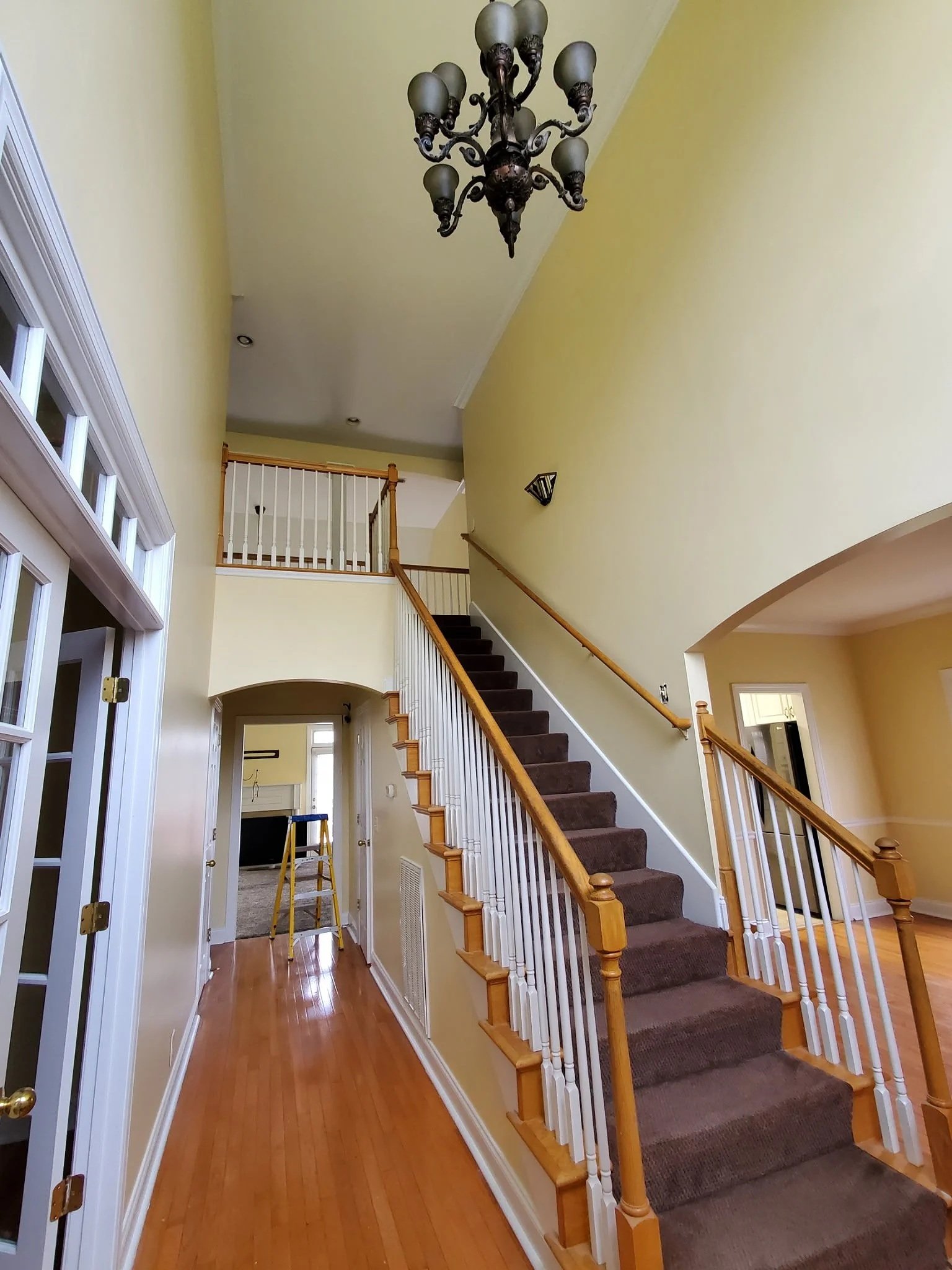 Interior view of a house staircase with wooden handrails, white spindles, and brown carpeted steps, with yellow walls, a chandelier hanging from the ceiling, and a hallway with windows and a ladder.