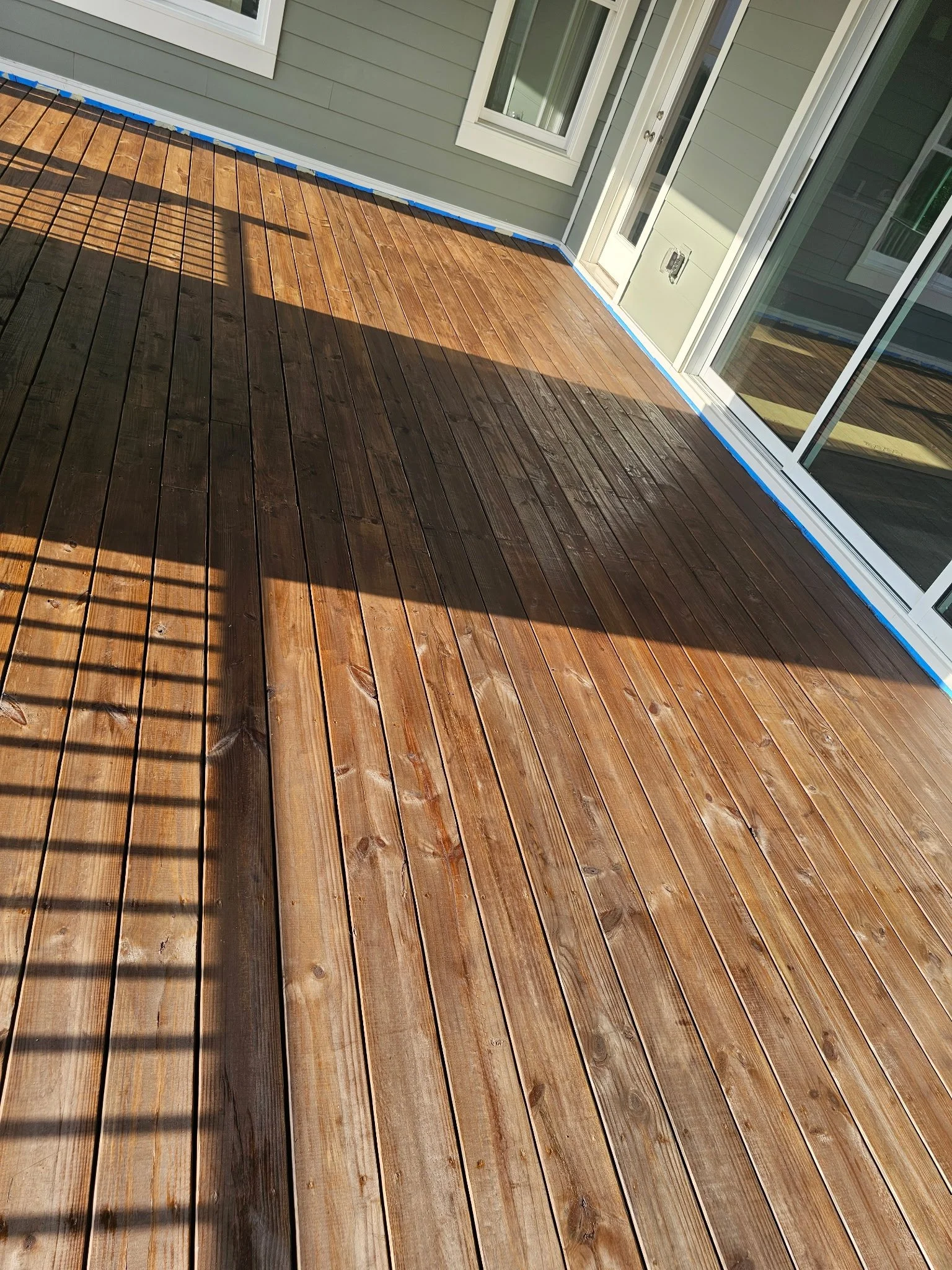 A wooden deck outside a house with sliding glass doors and windows, with sunlight and shadows cast on the deck.