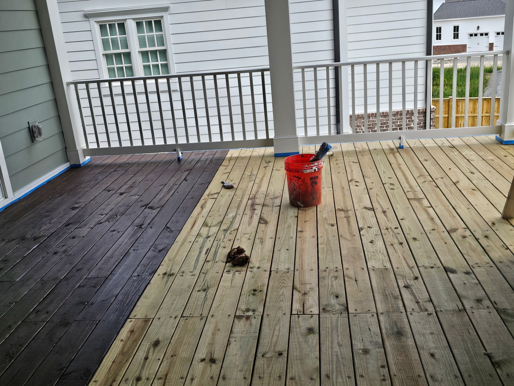 A porch with black and natural wood flooring, white siding, and a white railing, with a red bucket, a paintbrush, and a rag on the floor.