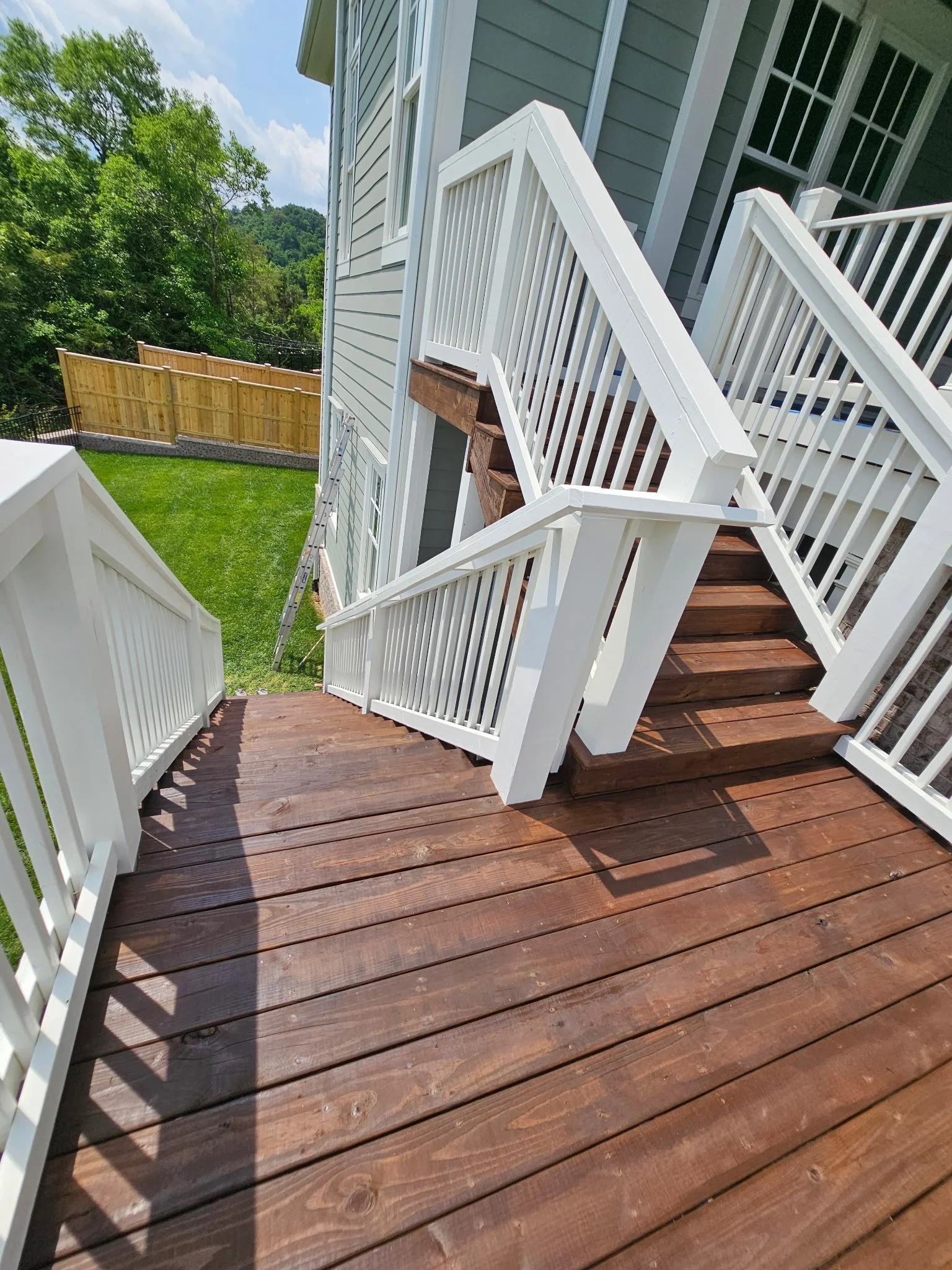 A wooden staircase with white railings leading down to a backyard with a green lawn and a wooden fence, with trees and a partly cloudy sky in the background.