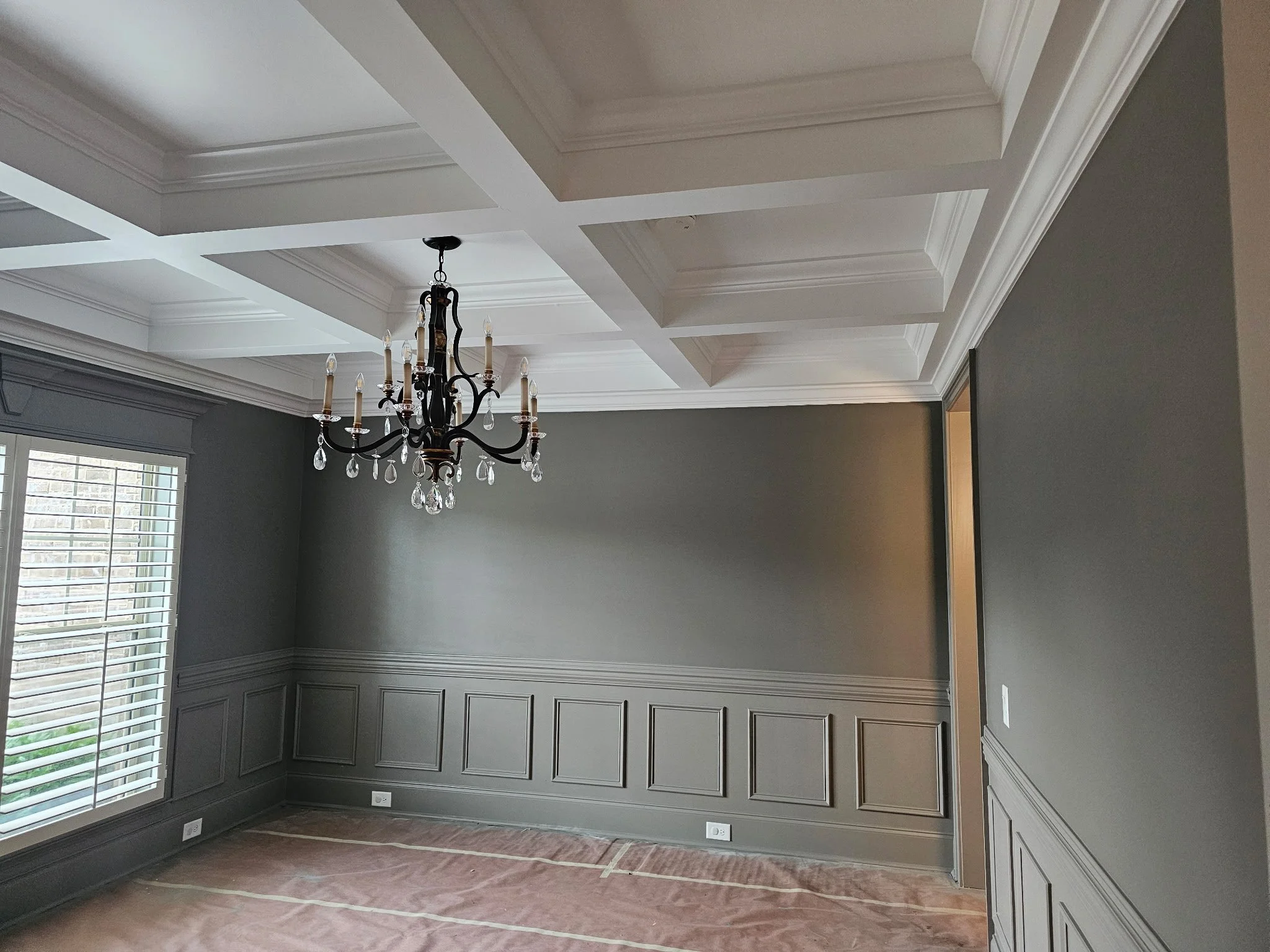 A room under renovation with painted gray walls, white wainscoting, a decorative ceiling with crown molding, a chandelier hanging from the ceiling, window with white blinds, and taped plastic on the floor.