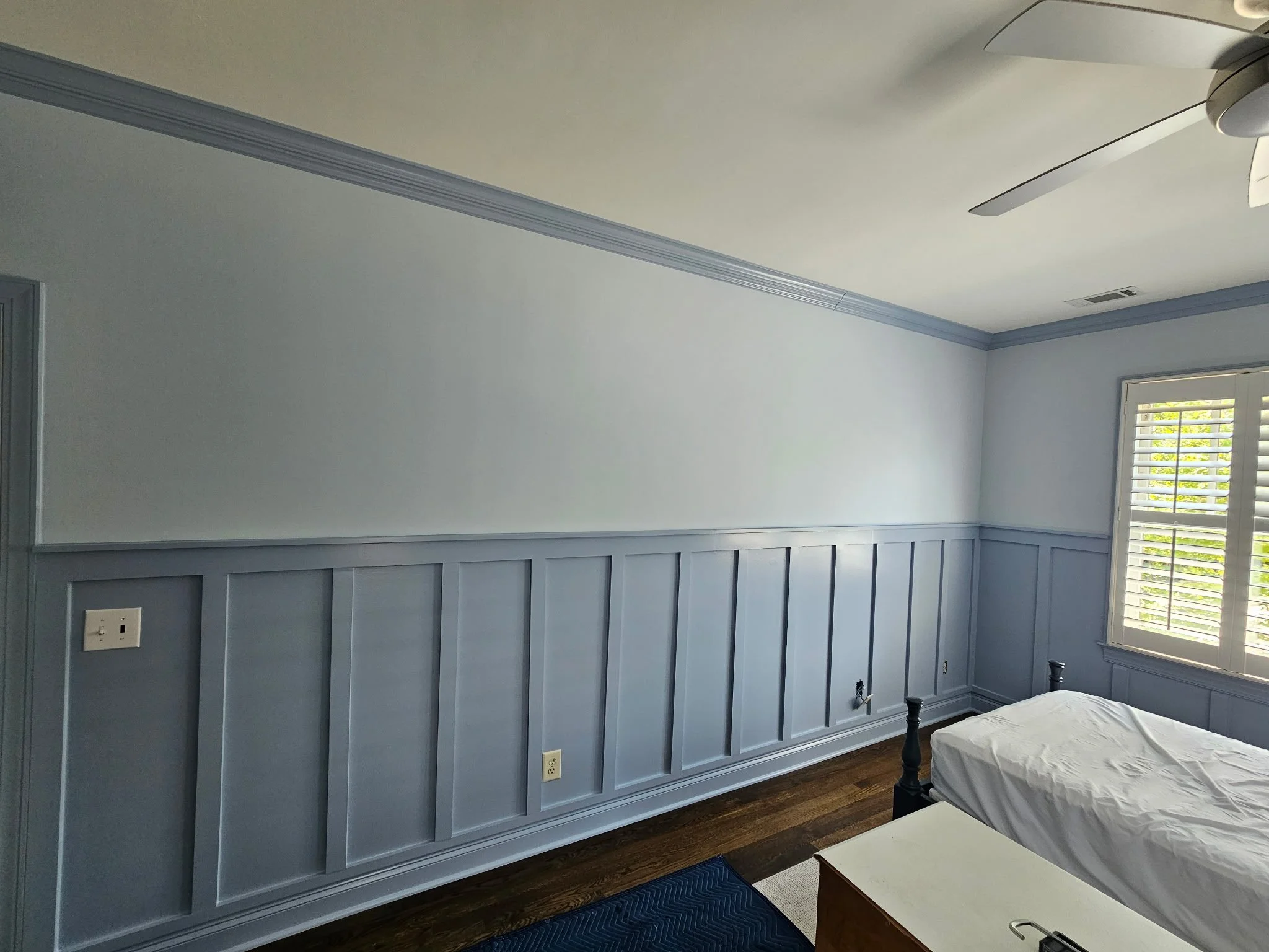 A bedroom with a light gray wall paneling, blue trim, and a large window with white blinds. Part of a bed with white bedding and a nightstand are visible, with wooden flooring.