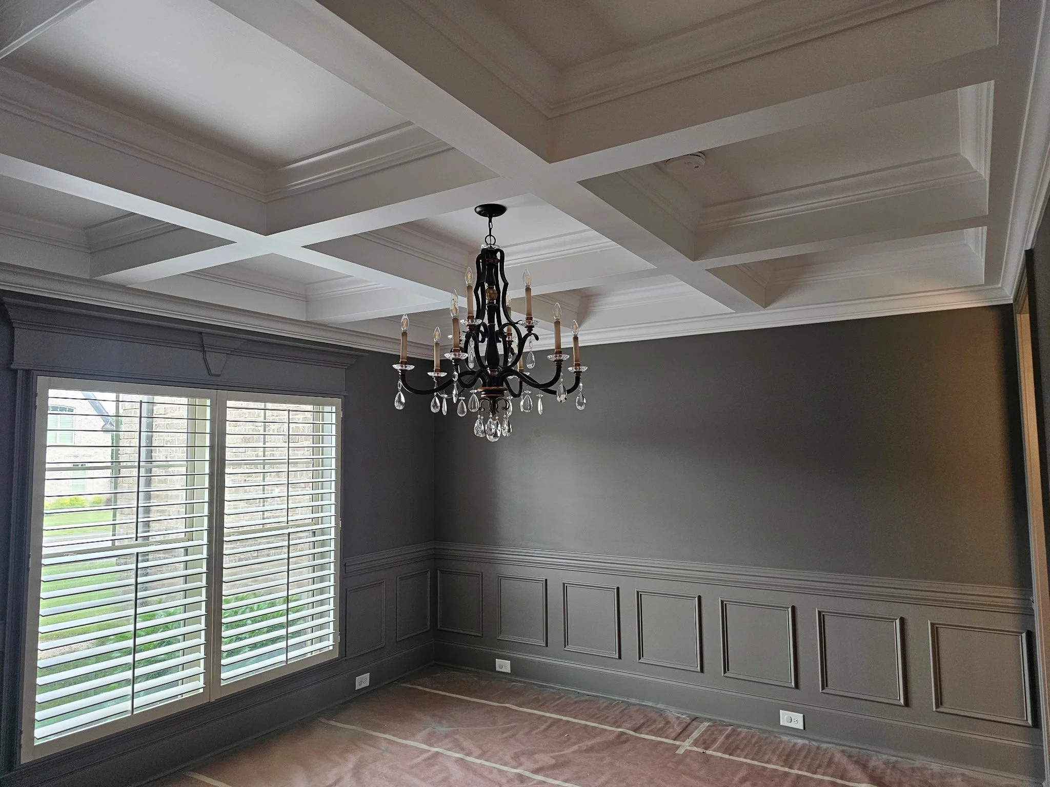 Interior of a room with gray paneled walls, large windows with white shutters, a chandelier hanging from a coffered ceiling, and electrical outlets near the floor.