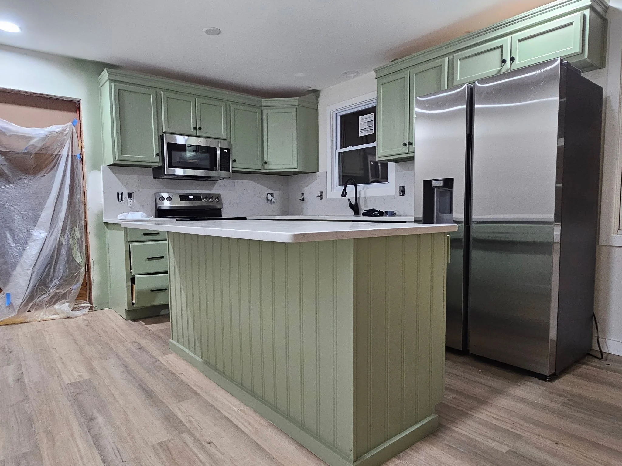 Kitchen with light green cabinets, stainless steel refrigerator, microwave, and stove. Contains a island with matching green cabinetry, a window, and visible construction materials cover part of the doorway.