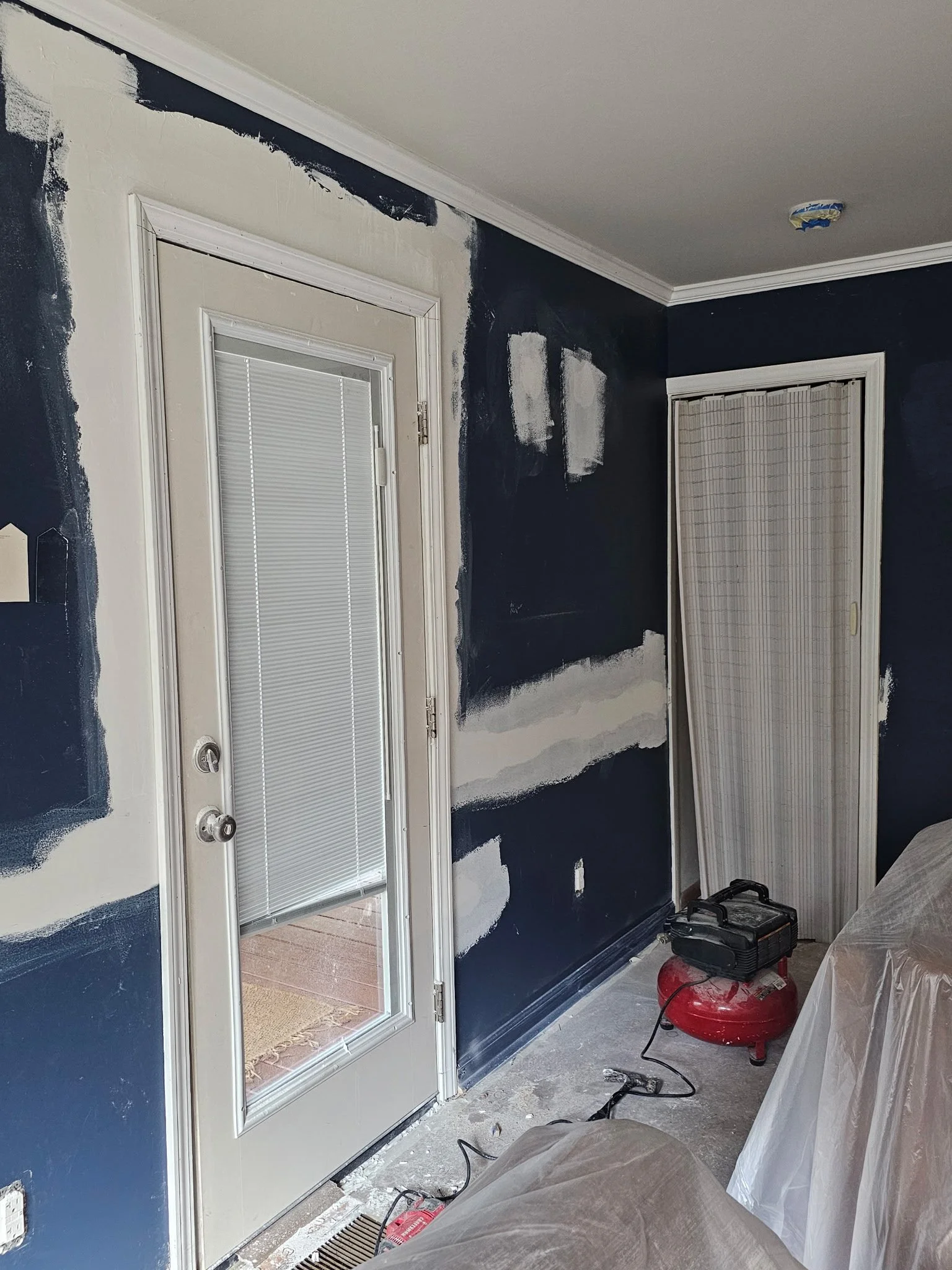 Room under renovation with dark blue walls, white crown molding, door with blinds, and construction tools including a red air compressor and a black toolbox.