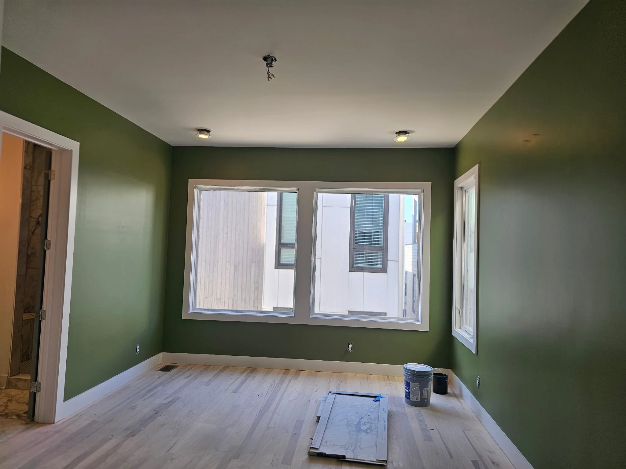 Interior of a room with green walls, two large windows, and unfinished ceiling with an electrical fixture. Painting supplies and tools are on the light wood floor.