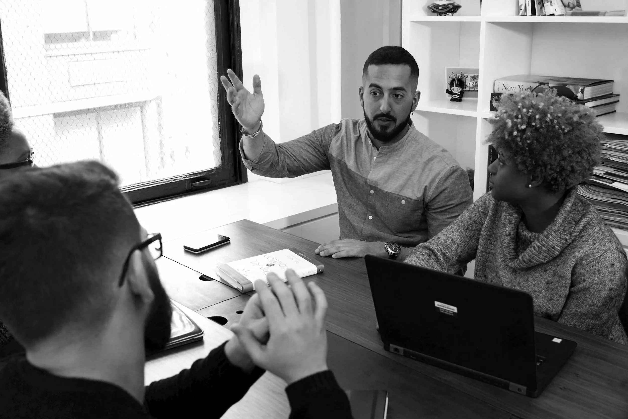 A man with a beard and short hair appears to be explaining something to a group of people in a meeting room. He is gesturing with his right hand. The room has a window with mesh, a bookshelf with books and decorative items, and a table with laptops and notebooks.