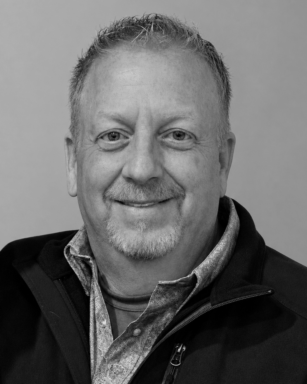 Black and white portrait of a smiling middle-aged man with short hair, light beard, wearing a patterned shirt under a zipped jacket.