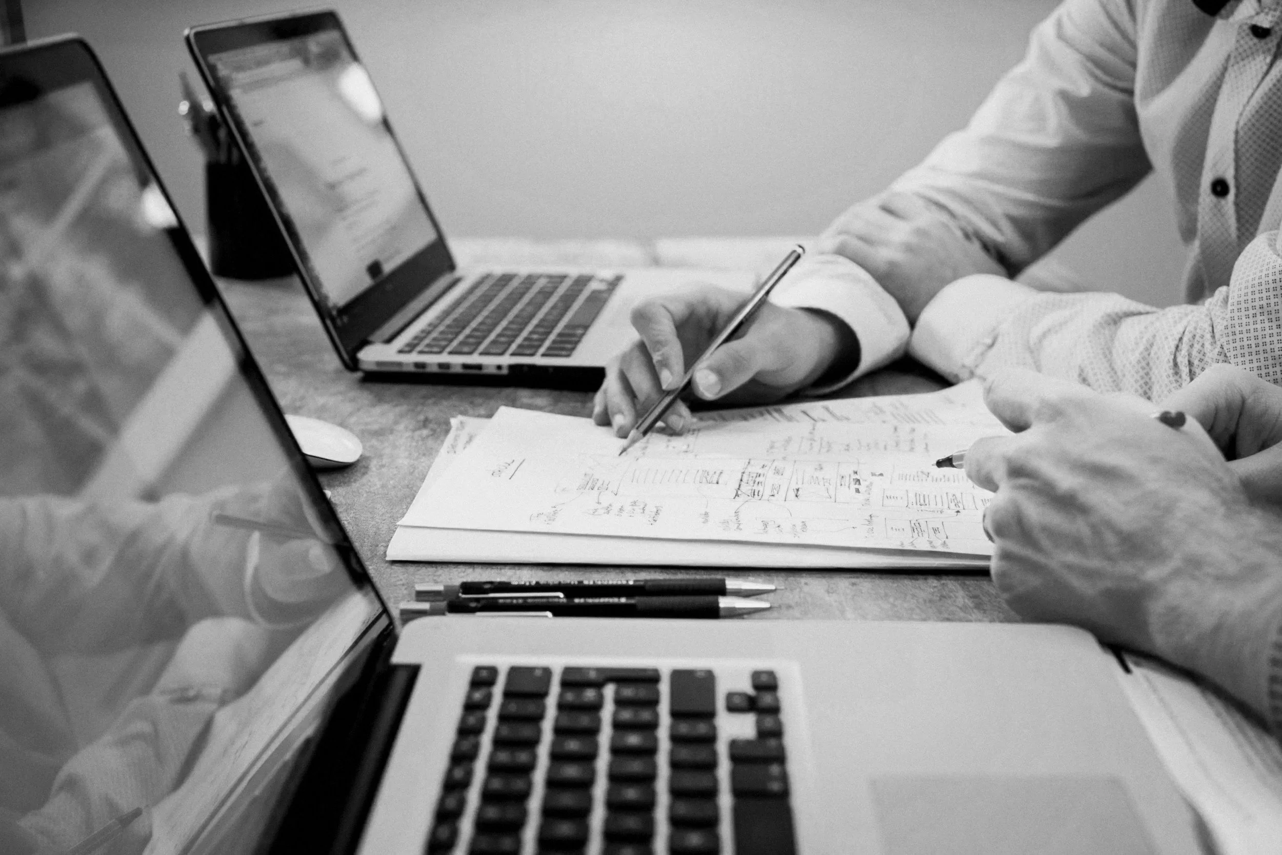 Black and white photograph of a person working at a desk, writing on papers with a pen, surrounded by open laptops and pens.