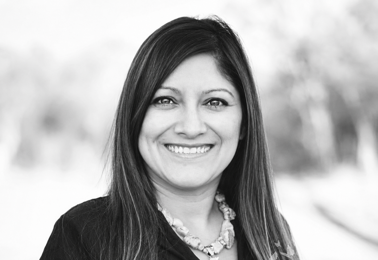 Black and white portrait of a smiling woman with long dark hair, wearing a necklace and a dark top, outdoors with blurred background.