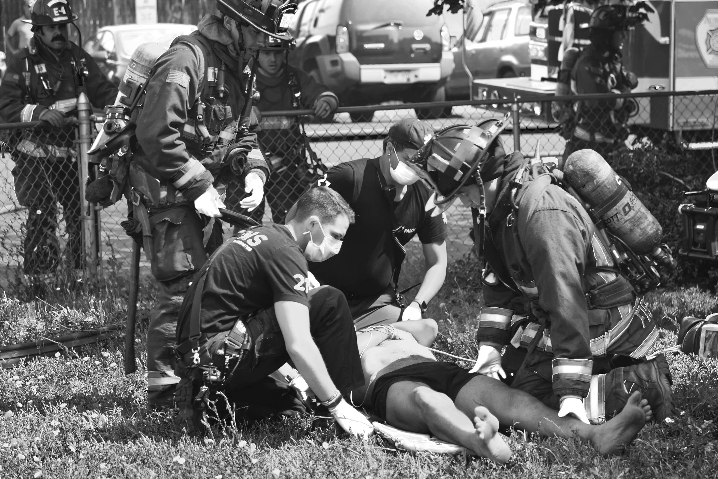 Firefighters performing rescue or medical assistance on a person lying on the ground, with others standing nearby behind a fence, in an outdoor setting.