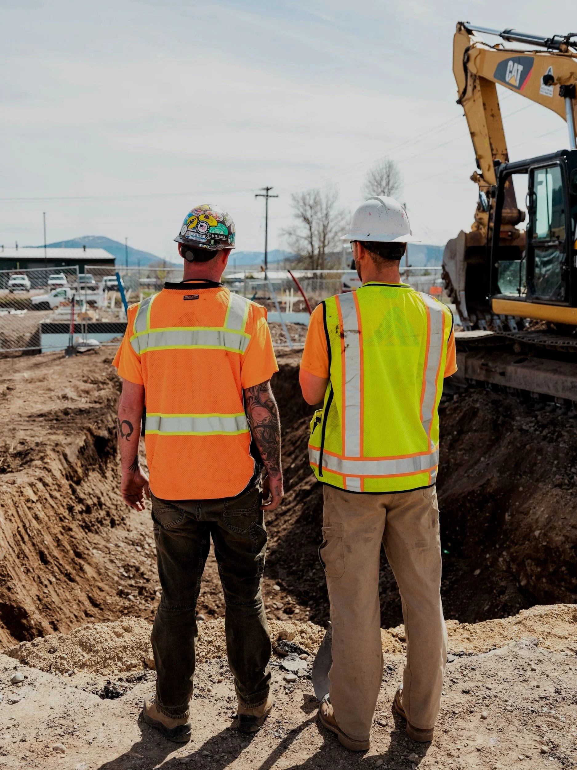 Two construction workers wearing safety vests and helmets standing near a large excavator at a construction site with mountains in the background.