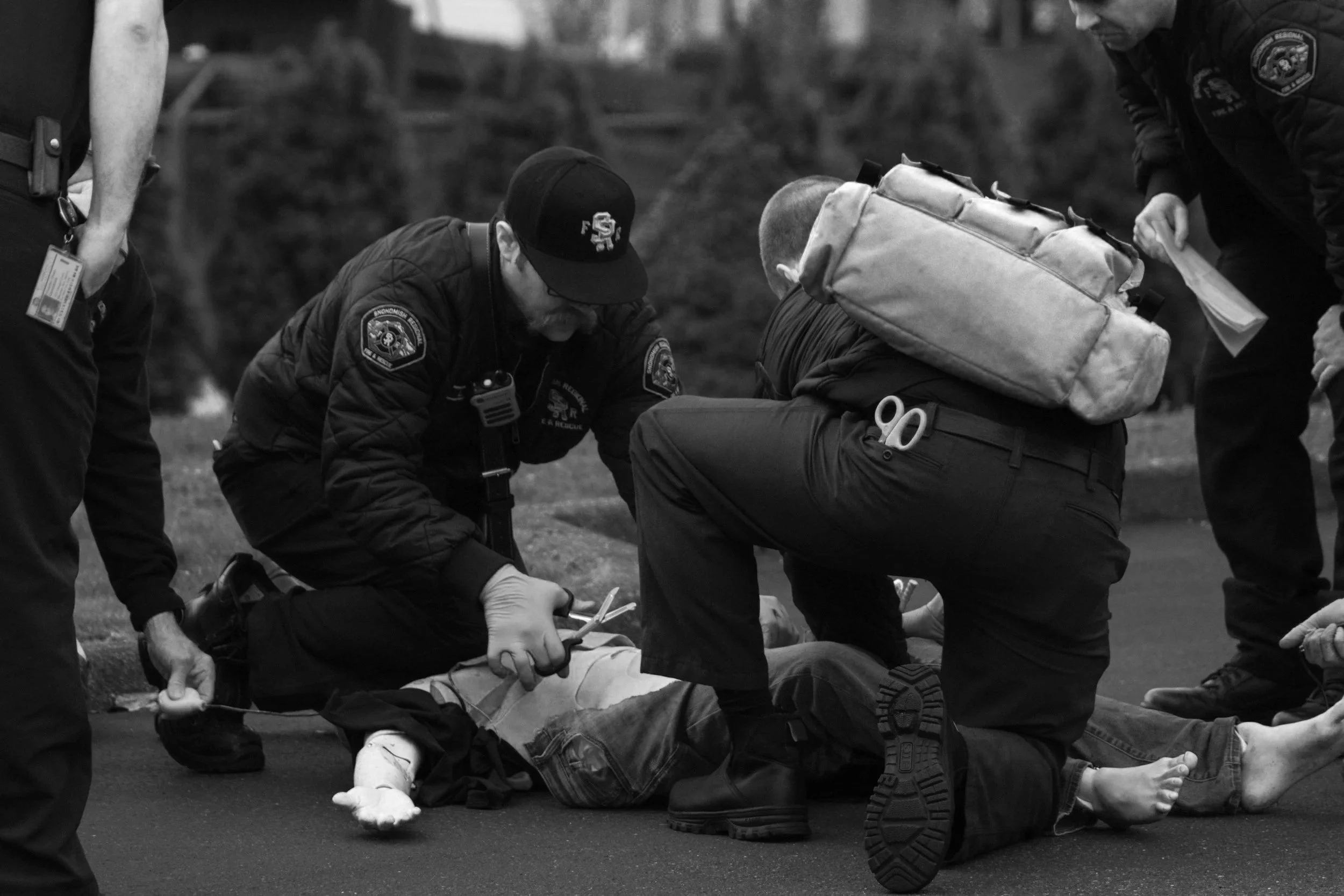 Black and white photo of emergency responders attending to a person lying on the ground, providing medical assistance with a padded splint and wearing uniforms with patches.