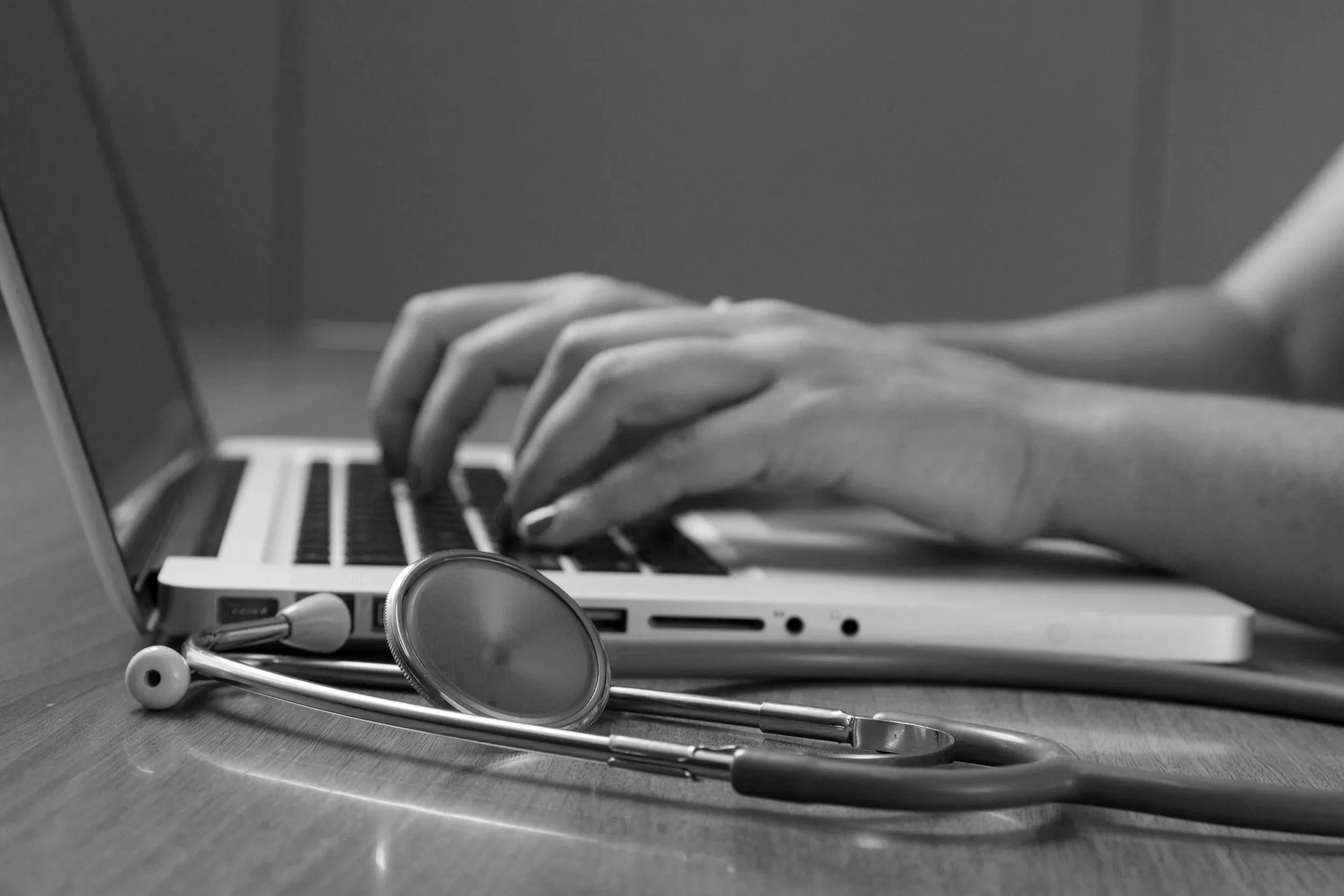 A stethoscope lying on a wooden surface in front of a laptop, with a stethoscope's earpieces and tubing visible, and the laptop screen reflecting the scene.