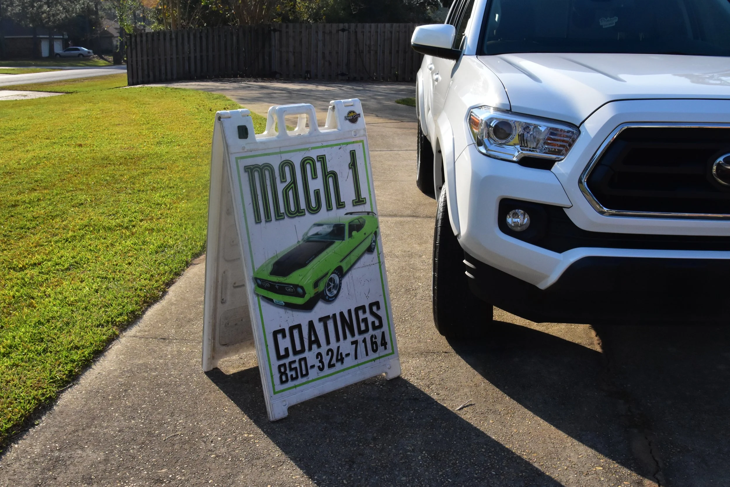 White SUV parked on driveway beside a sandwich board sign advertising car coatings with a green sports car illustration and contact number.