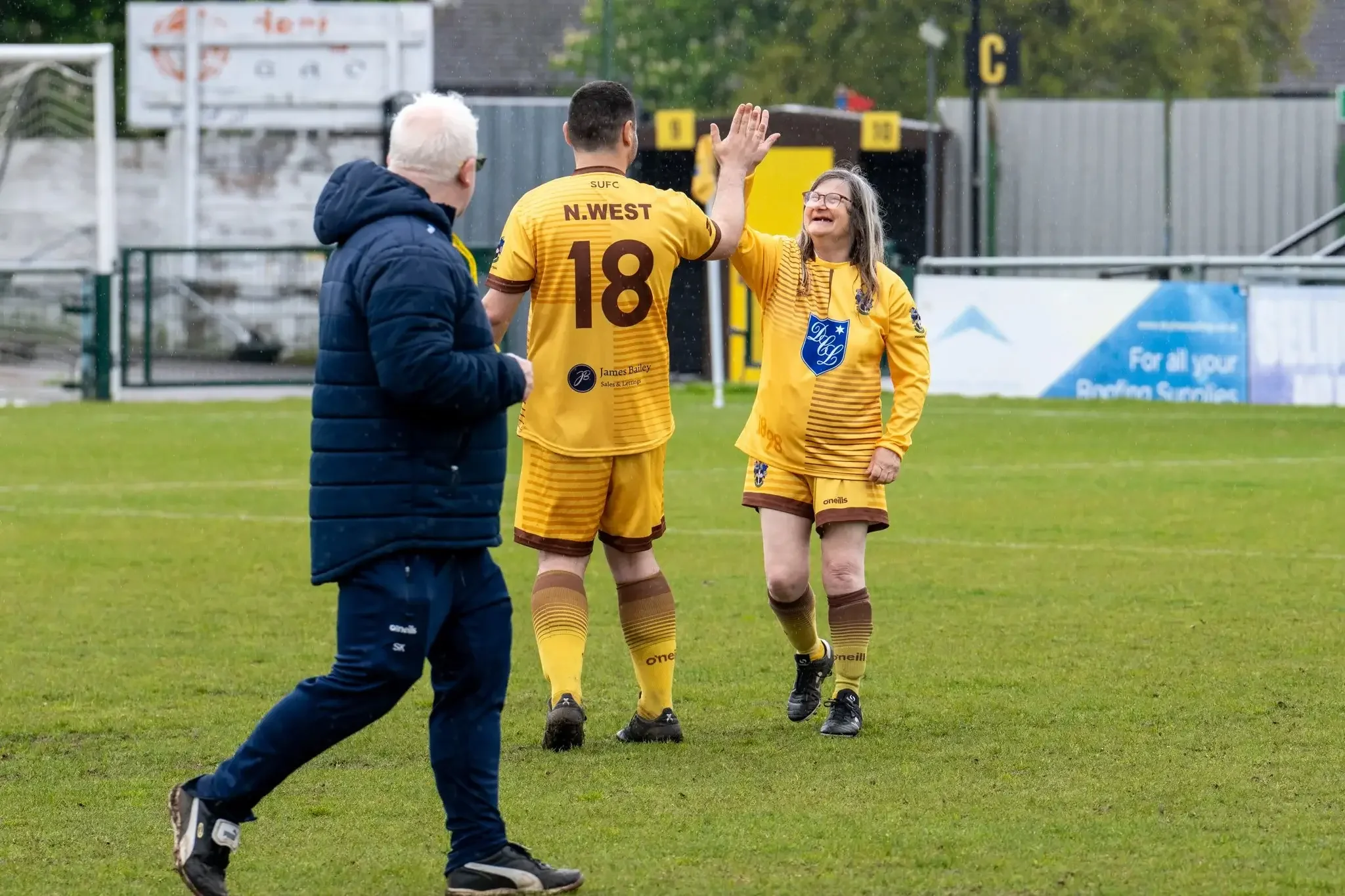 A soccer player in a yellow uniform high-fives a smiling woman in a matching yellow team shirt on a soccer field, with a man in a blue jacket walking nearby. Wind is blowing, and the background includes a fence and signs.