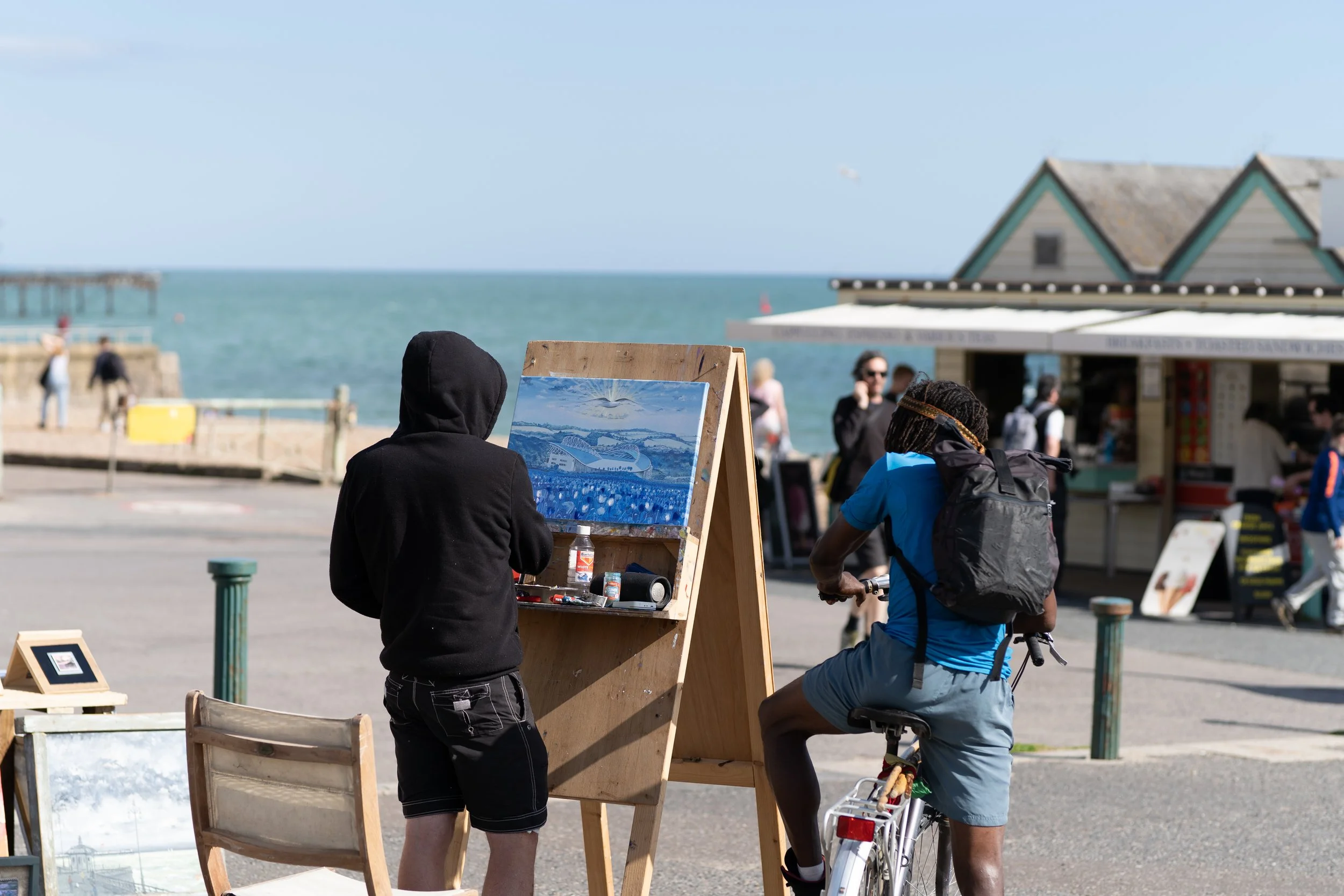 Beachside scene with an artist painting and a cyclist, people in the background near the water and pier, overcast sky.