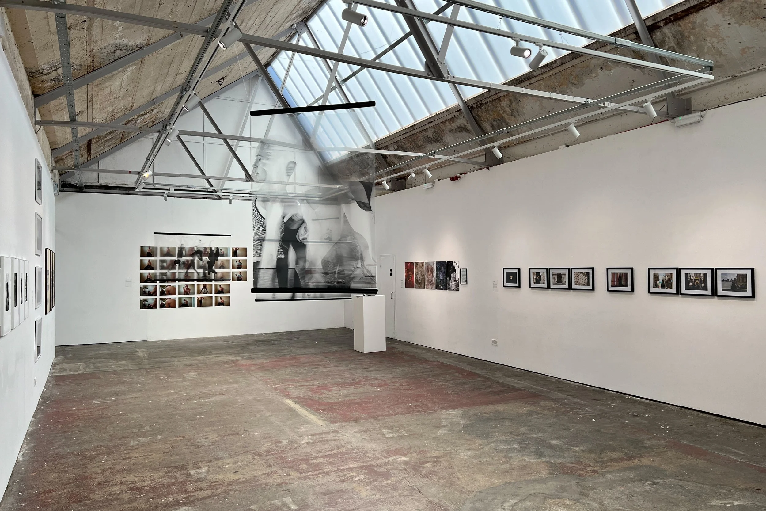 An art gallery with photos and artwork on white walls, a large black-and-white portrait of a woman on a hanging scroll, and a ceiling with exposed beams and skylights.