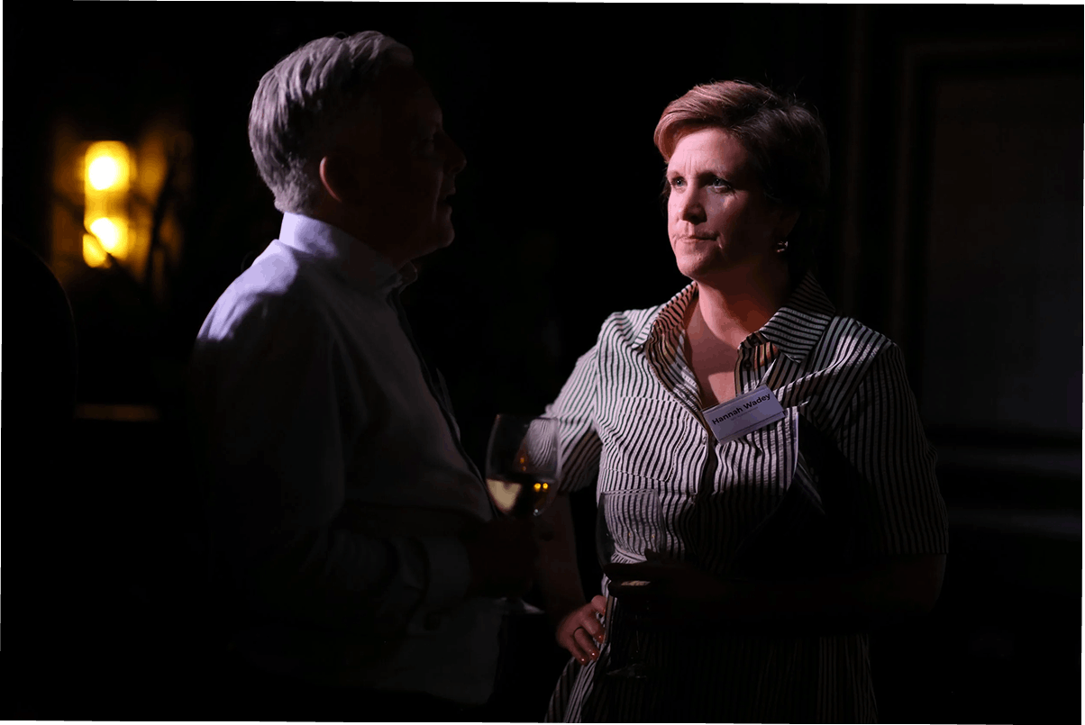 A man and a woman with serious expressions talking at a social event, holding wine glasses, in a dimly lit room with background lights.