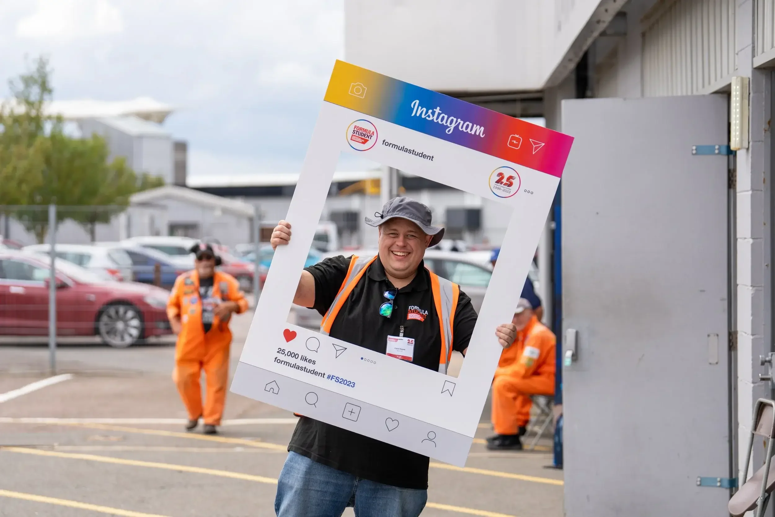 Man smiling and holding a large Instagram frame cutout at an event, with other people in orange safety vests in the background in a parking lot.