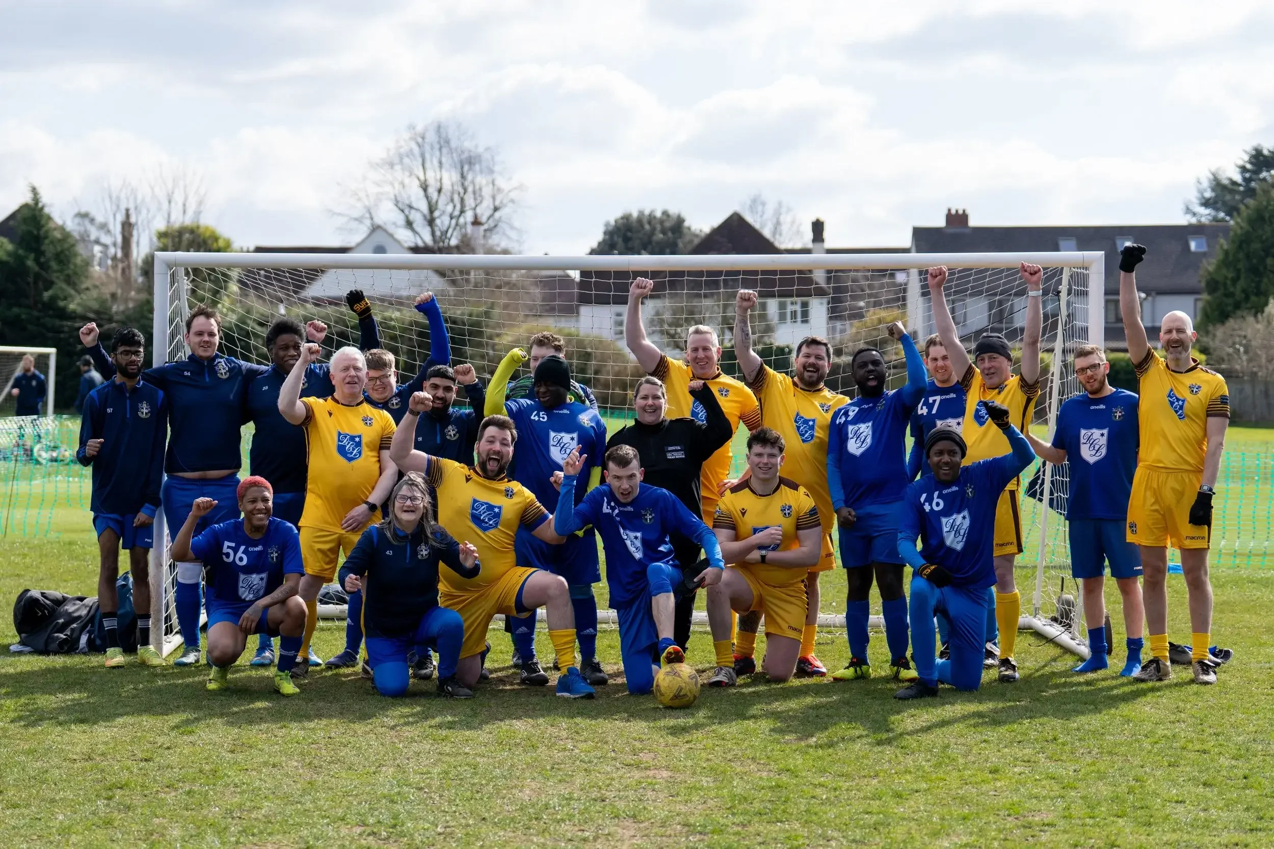 Group of soccer players and supporters celebrating on a grass field in front of a goalpost, with houses and trees in the background.