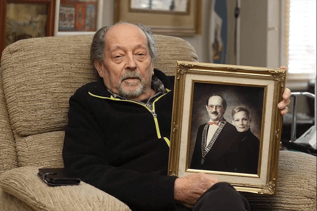 An elderly man sitting in a beige armchair holding a framed portrait of a man and a woman dressed formally.