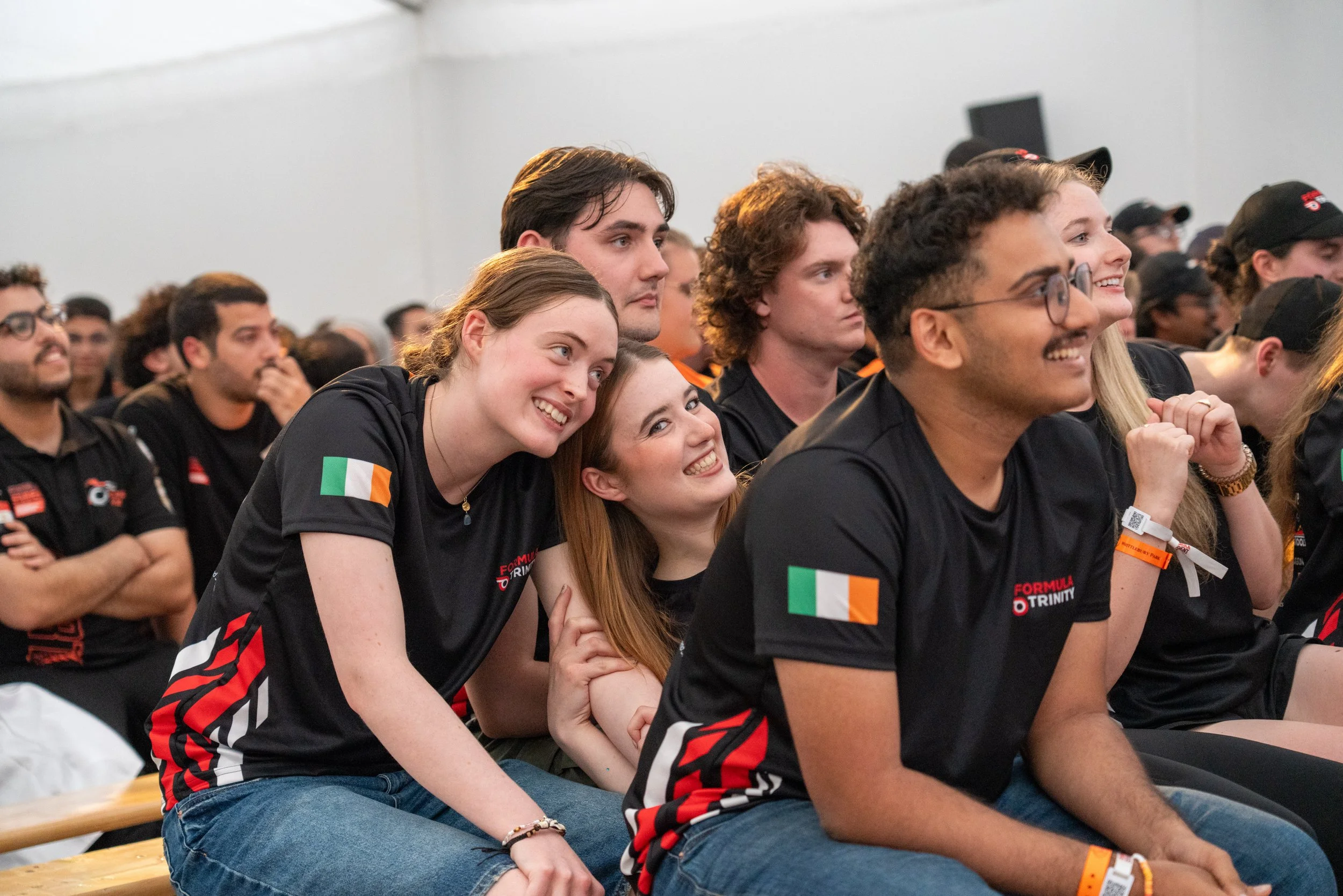 Group of young people sitting and smiling, attending an event, some wearing black shirts with Italian flag patches, in a large indoor space.