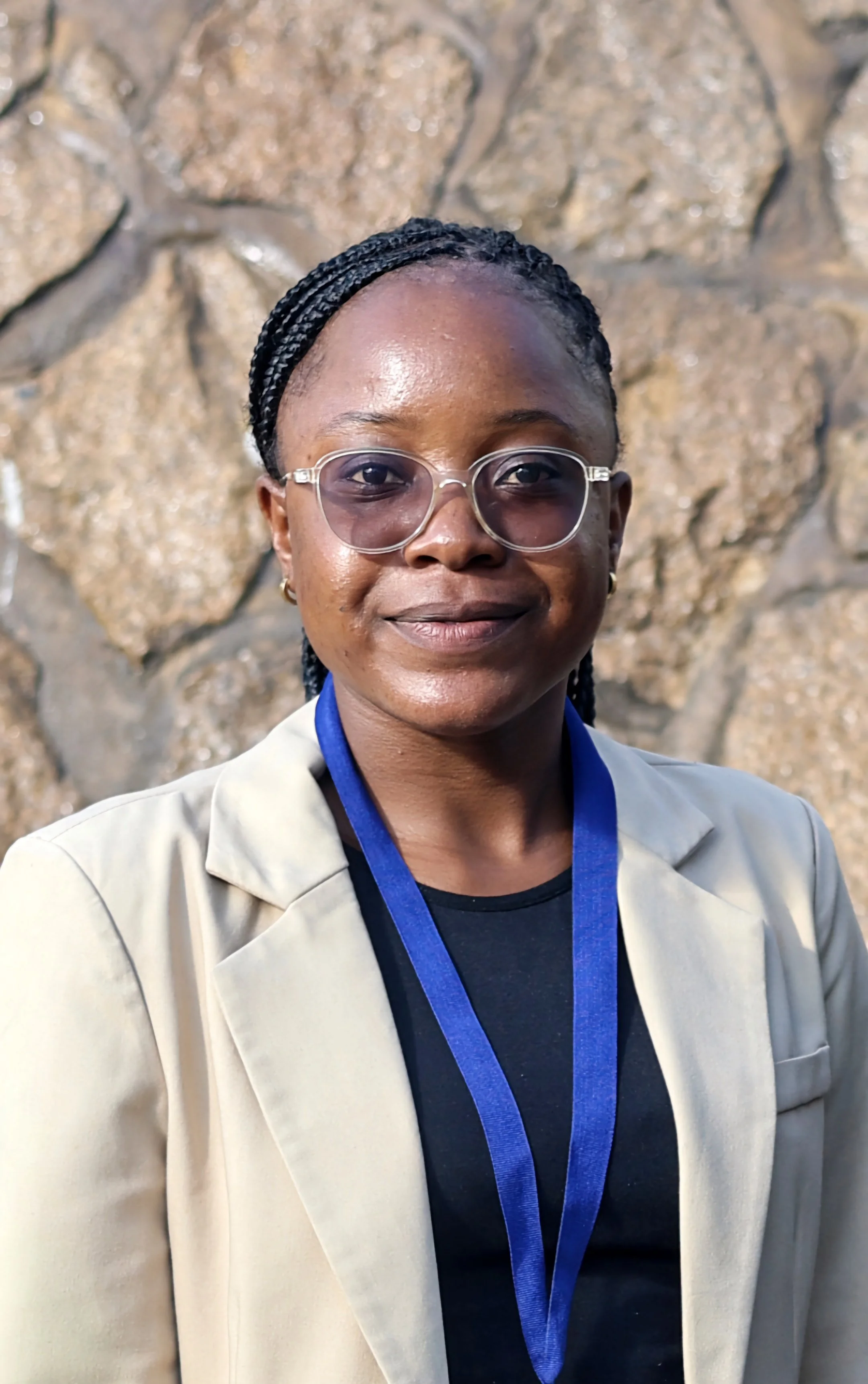 Close-up of a woman wearing glasses, a beige blazer, and a lanyard, standing in front of a stone wall.
