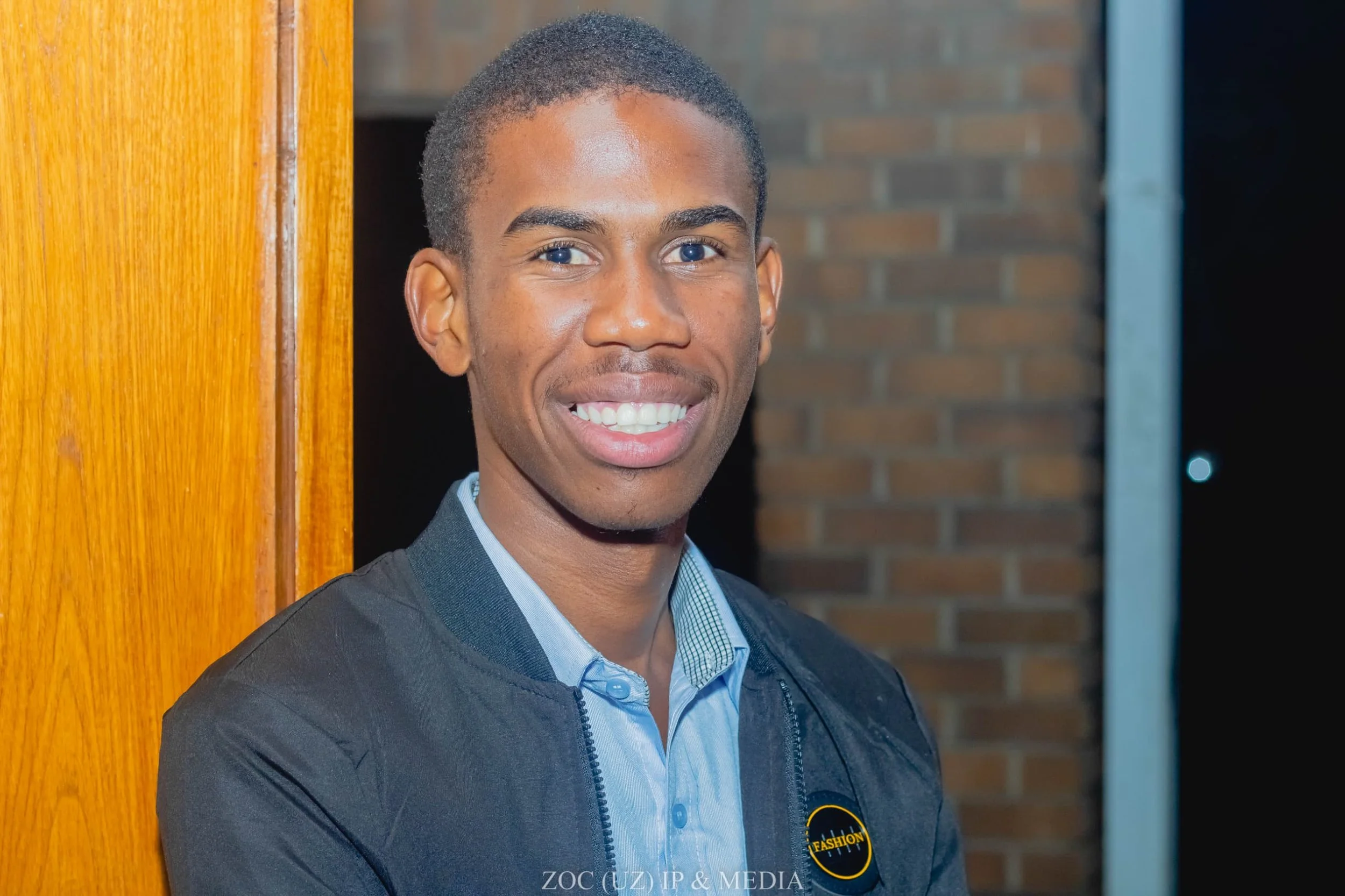 A young man with short black hair, blue eyes, and a bright smile, dressed in a blue shirt and dark jacket, is standing next to a wooden panel with a brick wall in the background.