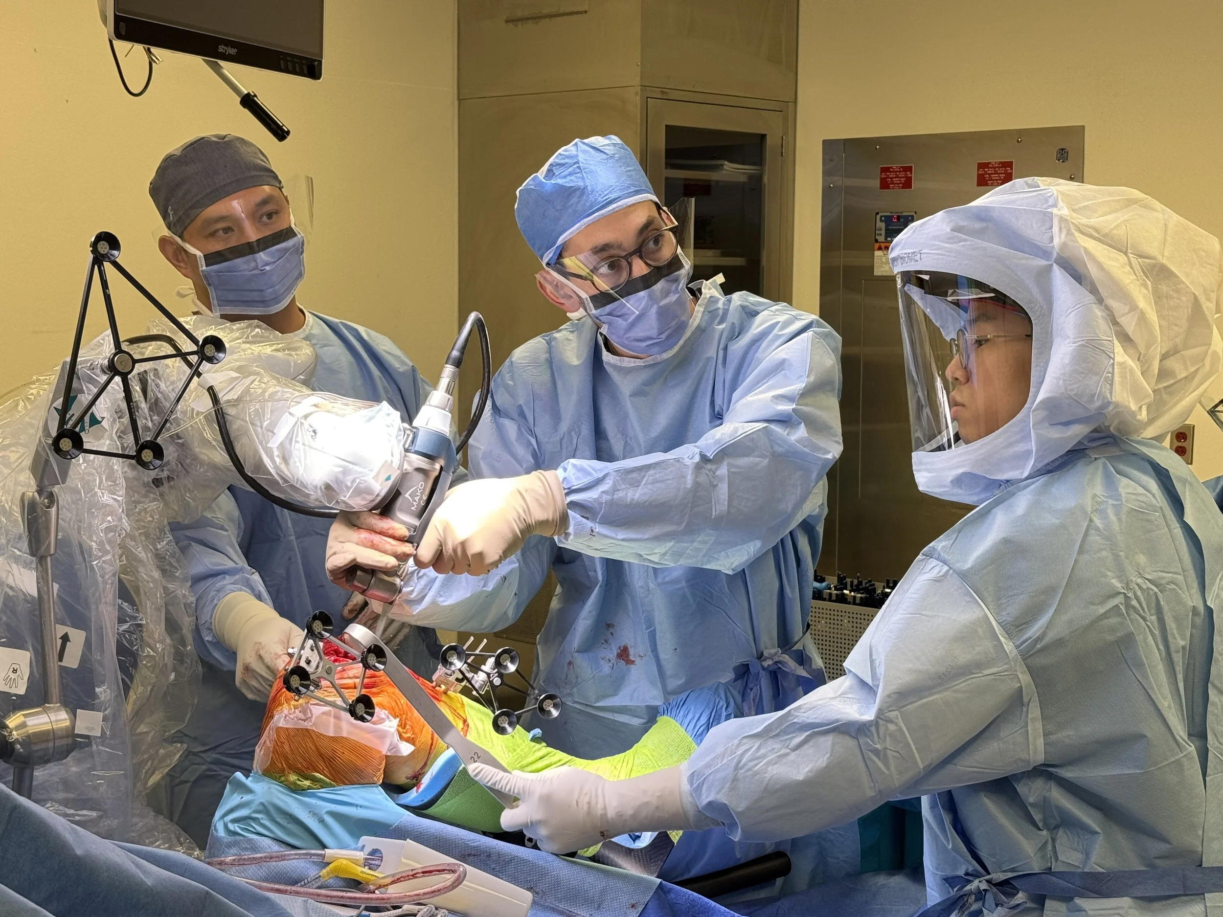 Medical team performing surgery in an operating room, with surgeons wearing blue scrubs, masks, and gloves, focused on a patient covered with sterile drapes.
