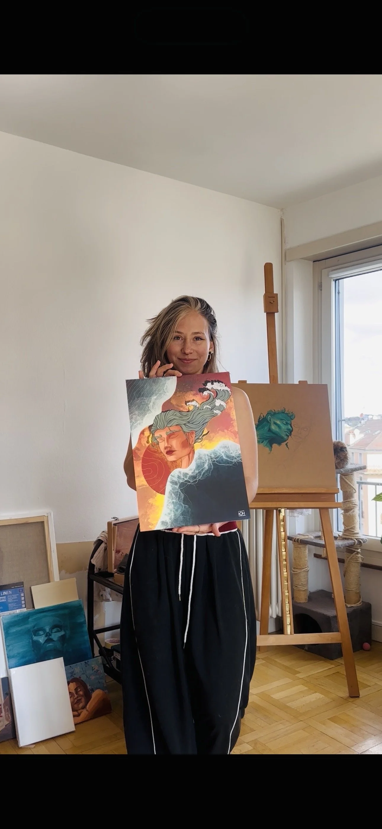 Une femme souriante tenant une peinture avec une femme aux cheveux de vagues et un fond vibrant, dans un studio d'art avec d'autres œuvres en arrière-plan.