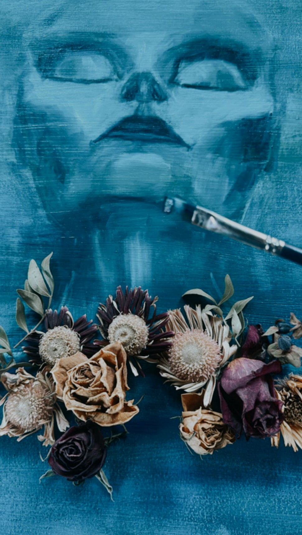 Une peinture d' visage en gros plan. Au premier plan, un pinceau et un arrangement floral composé de roses, d'eucalyptus et d'autres fleurs séchées.