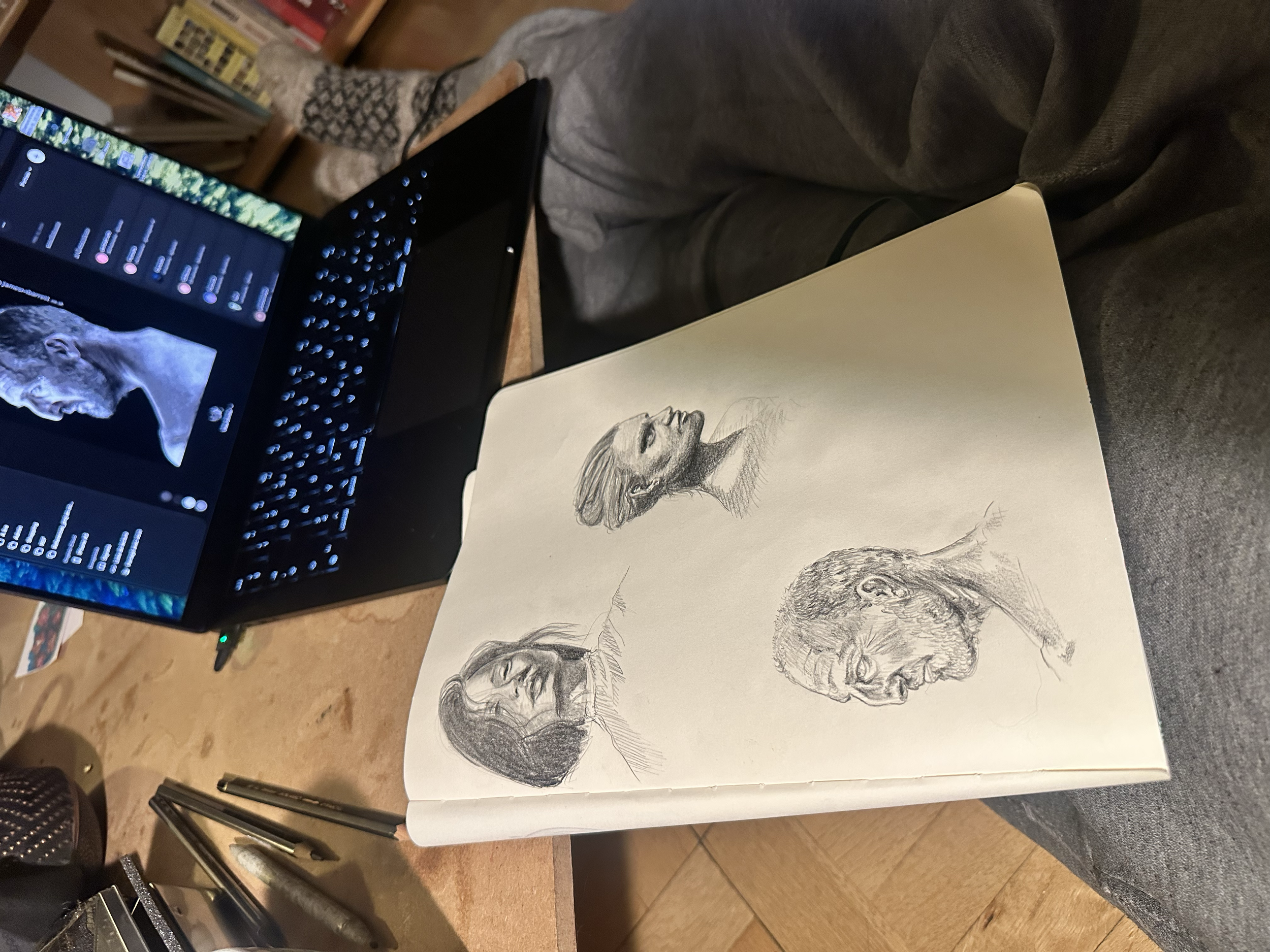 Croquis de plusieurs portraits de visages masculins et féminins en techniques de dessin différentes, déposés sur une table en bois, avec un ordinateur portable à gauche.