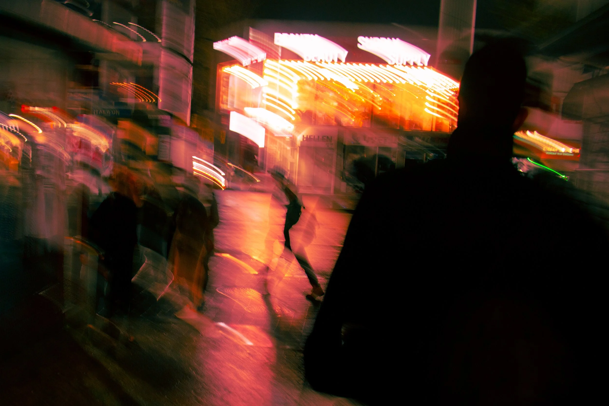 Une scène urbaine nocturne avec des lumières de néon floues, silhouettes de personnes et de bâtiments, ambiance dynamique et colorée.