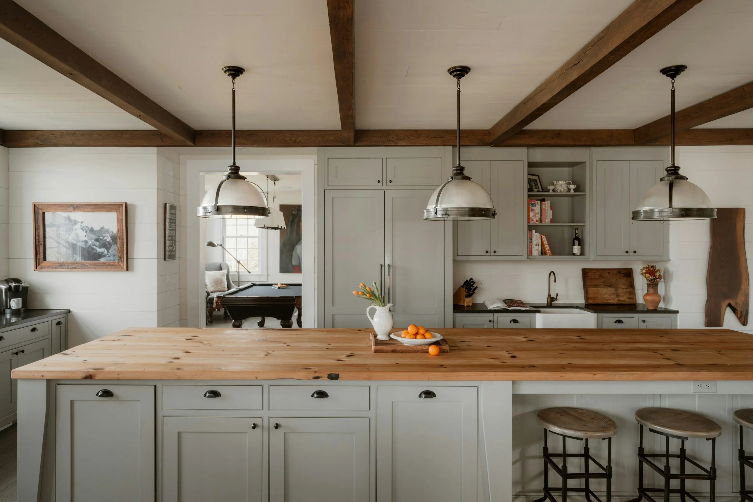 A spacious kitchen with a large wooden island, gray cabinets, white walls, and pendant lights hanging from a wooden beam ceiling. There is a bowl of oranges and a white pitcher on the island, and a photo frame and open shelves with dishes and books on the wall.