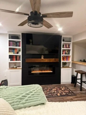 Living room with a wall-mounted TV above an electric fireplace, white built-in shelves on both sides filled with books, a ceiling fan with wooden blades, a green blanket on a light-colored sofa, and a small rug on dark hardwood flooring.