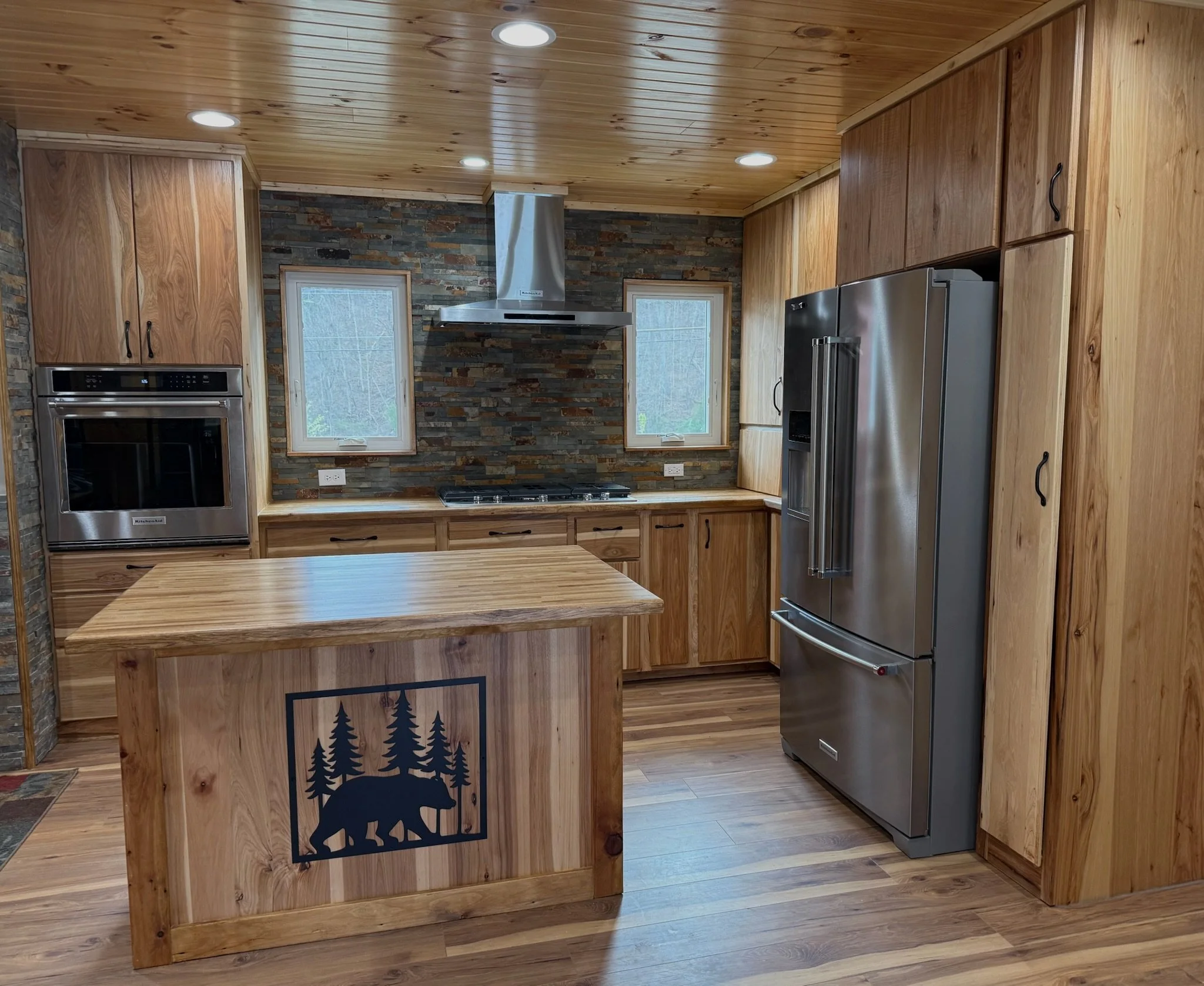 A rustic kitchen with wooden cabinets, a stainless steel refrigerator, and a stone accent wall. There are two small windows above the stove, with a range hood between them. A wooden kitchen island with a bear and trees engraved on the front is also visible.