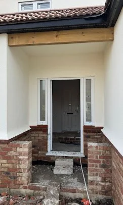 Front entrance of a house under construction with a black door and brick steps, partially built with unfinished concrete and surrounding white walls.