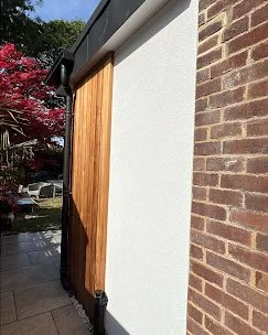 A wooden sliding door set into a white wall beside a brick wall, with a garden and parked cars in the background.